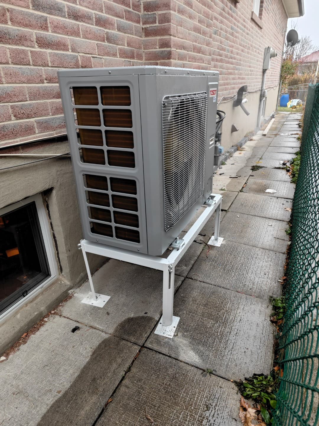 Side view of a gray outdoor heat pump on a stand along a concrete walkway