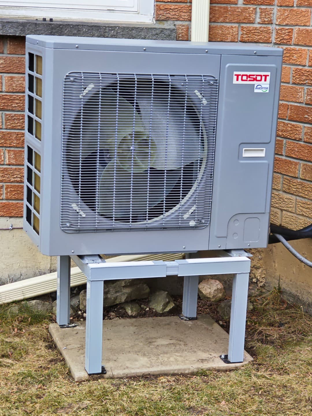 Front view of a gray outdoor heat pump on a stand in front of a brick wall