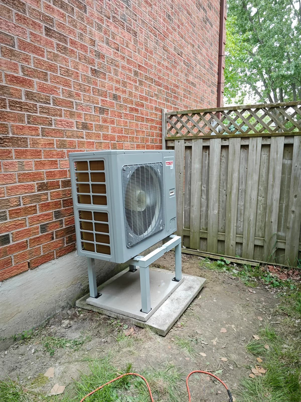 Gray outdoor heat pump on a stand beside a brick wall and a wooden fence