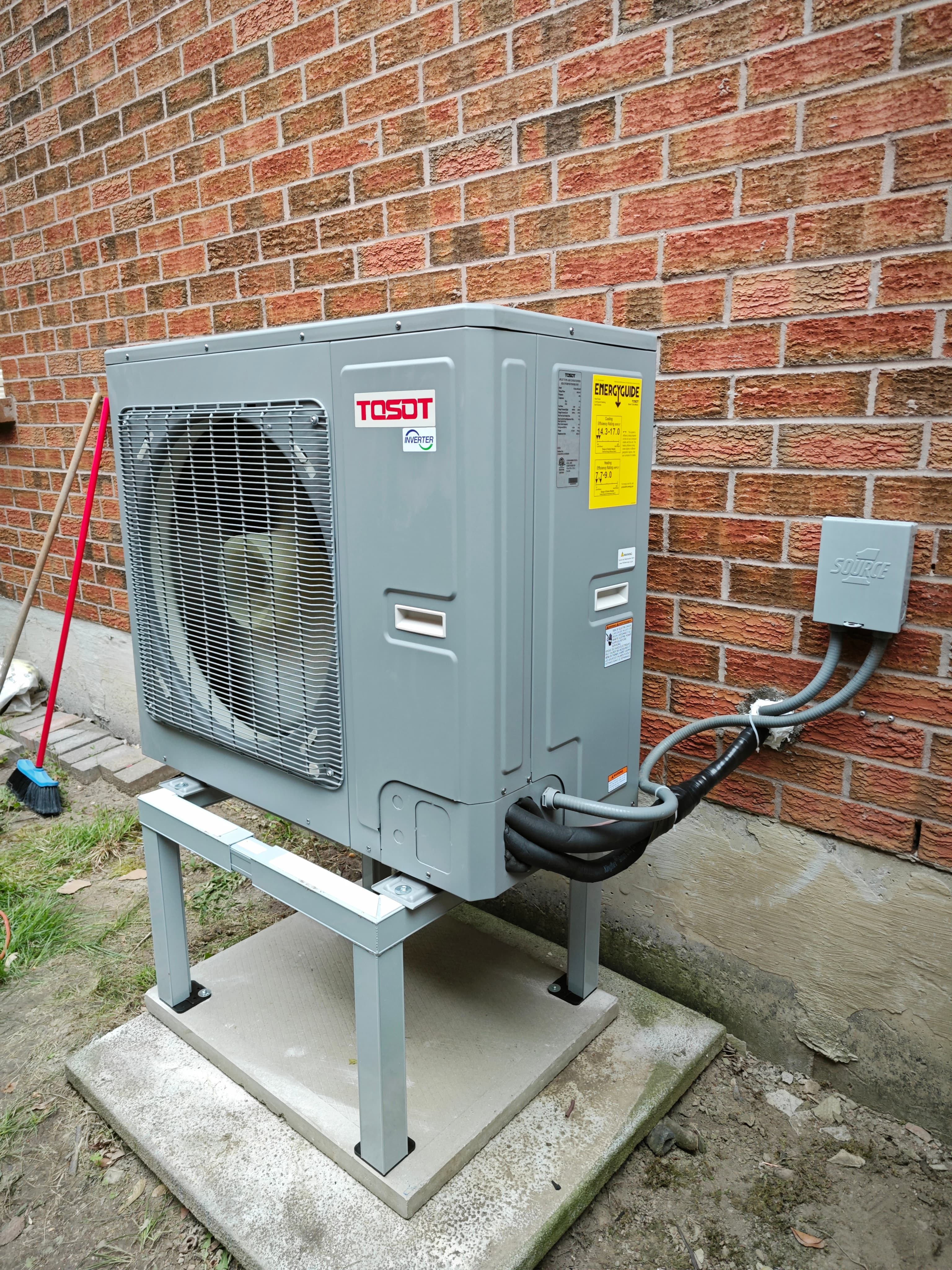 Gray outdoor heat pump on a metal stand, with refrigerant lines and a wall disconnect